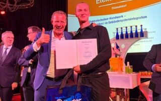 Zwei Personen auf der Bühne der Elektro-Innung Düsseldorf halten eine Abschlussurkunde in die Kamera und lächeln, im Hintergrund ist eine Präsentationsfolie zu sehen.