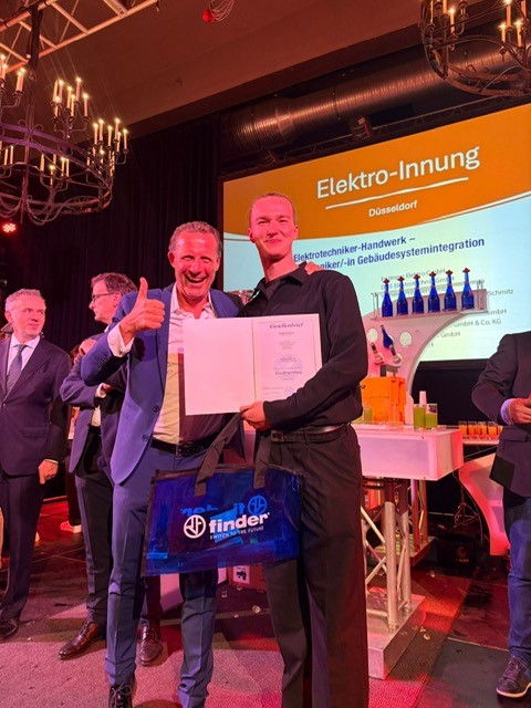 Lossprechung Bester Azubi Zwei Personen auf der Bühne der Elektro-Innung Düsseldorf halten eine Abschlussurkunde in die Kamera und lächeln, im Hintergrund ist eine Präsentationsfolie zu sehen.