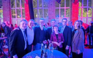 Zwei Personen auf der Bühne der Elektro-Innung Düsseldorf halten eine Abschlussurkunde in die Kamera und lächeln, im Hintergrund ist eine Präsentationsfolie zu sehen.